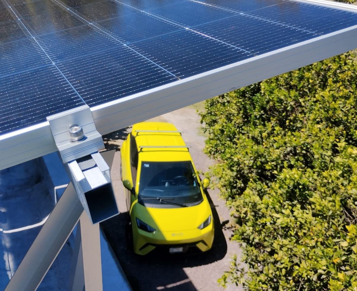 Yellow car parked under a solar panel carport with metal support beams and growing green shrubs nearby, viewed from above.